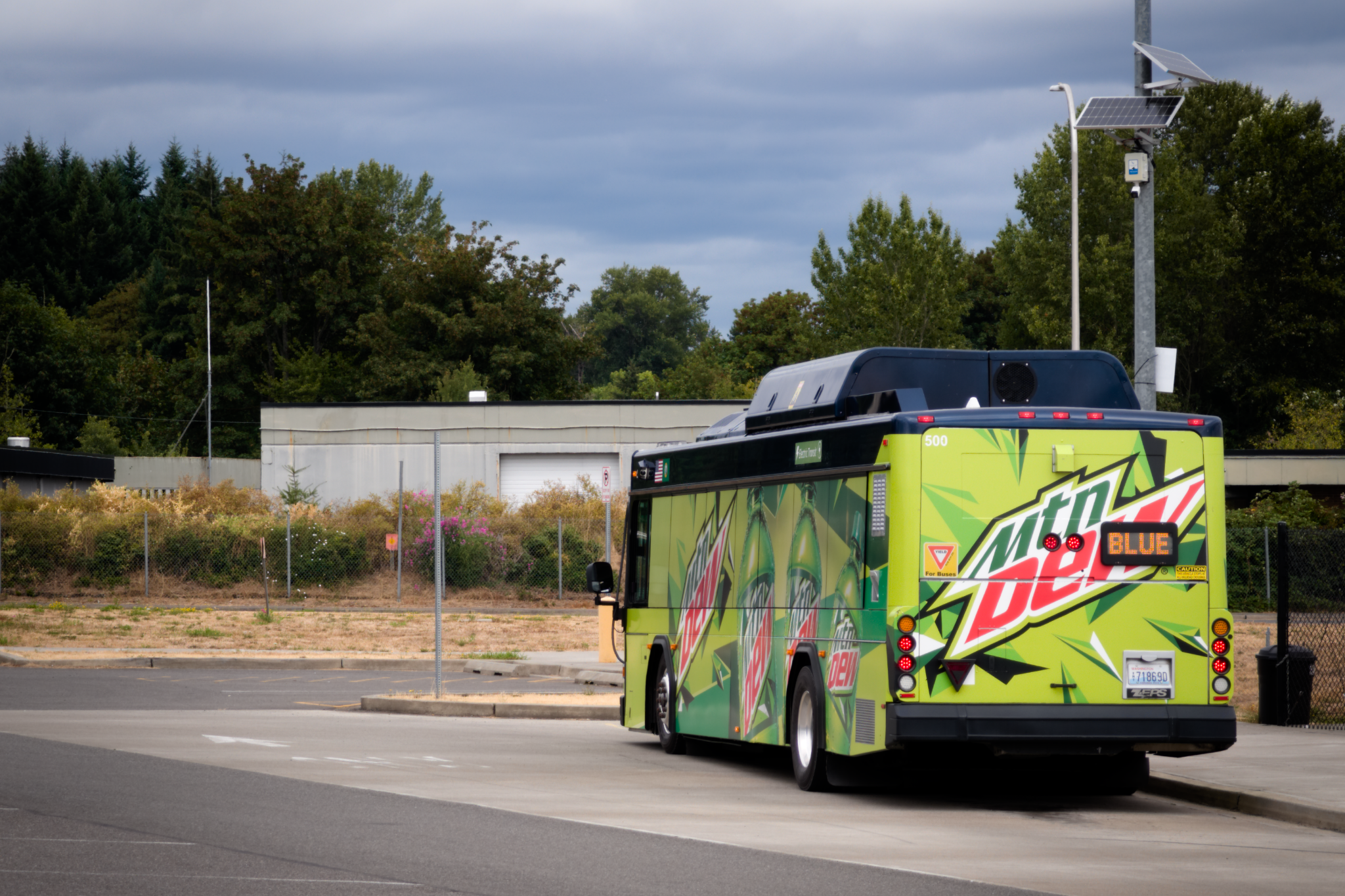 Photograph of Lewis County Transit 500, a 2004 Gillig Low Floor 35-foot bus converted by Complete Coach Works to a ZEPS bus. Seen here, the Gillig is wrapped in a full ad wrap for Mountain Dew, from dotted windows featuring depictions of the bottles to a large Mountain Dew logo on the rear of the bus. A rear headsign indicates the bus is operating on the Blue Line and is pictured laying over in a bay at the Centralia transit center, contrasting an overcast deep blue sky in the background. A large black bulge on the midsection of the roof can be seen with a yellow warning sticker and cooling fans, typical of ZEPS-converted buses, albeit quite abnormal compared to factory battery-electric Gilligs.